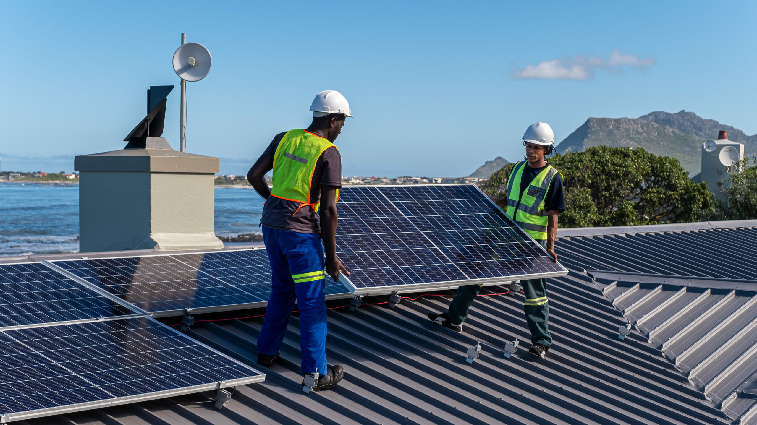 Two African men install solar panels neatly on a residential roof of a house near the ocean. Sustainable living.